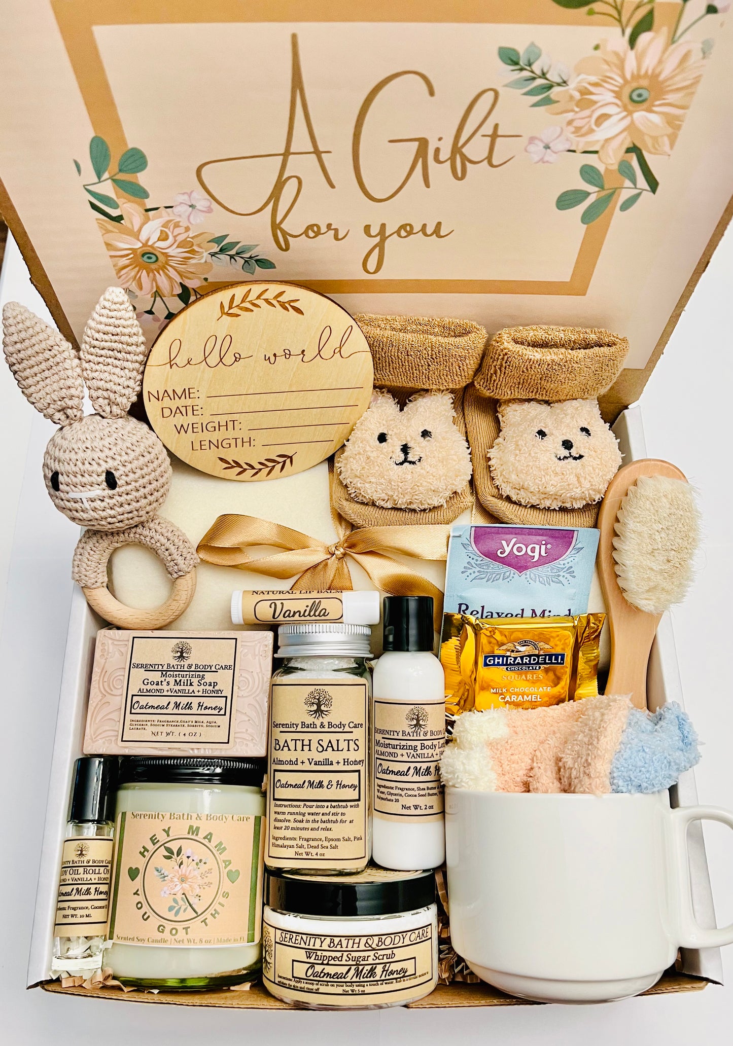 A gift box open to reveal various items including bath products, a mug, socks, a baby rattle, a bear-shaped sock, a wooden hair brush, and a Hello World announcement sign, all arranged neatly inside.